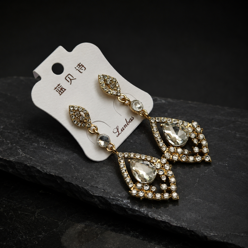 Pair of gold and crystal earrings on a black stone surface with a white card displaying brand name.